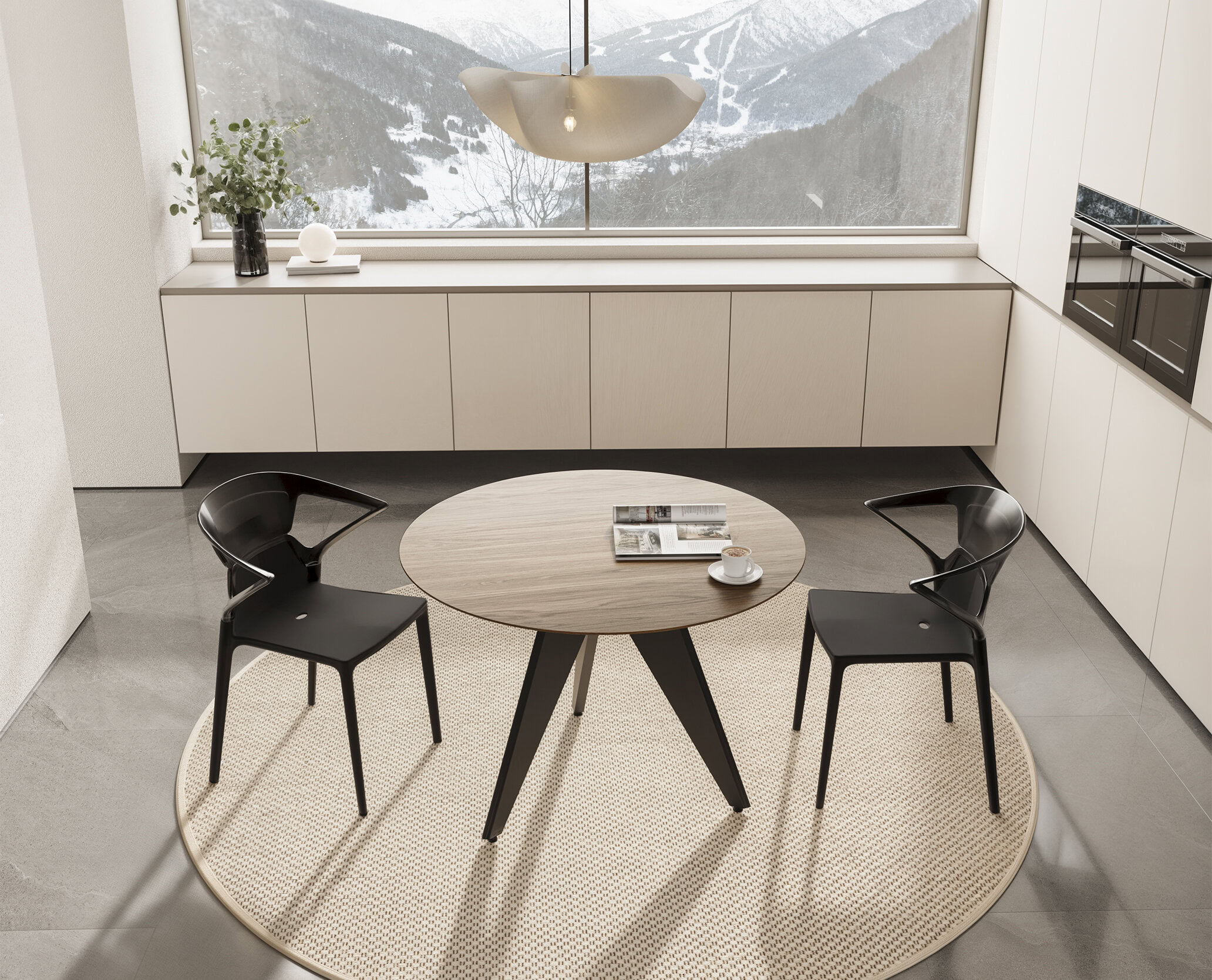 Contemporary round wooden dining table with two black modern chairs on a beige rug, bright minimalist kitchen design with white cabinetry and large window showcasing snowy mountain scenery.