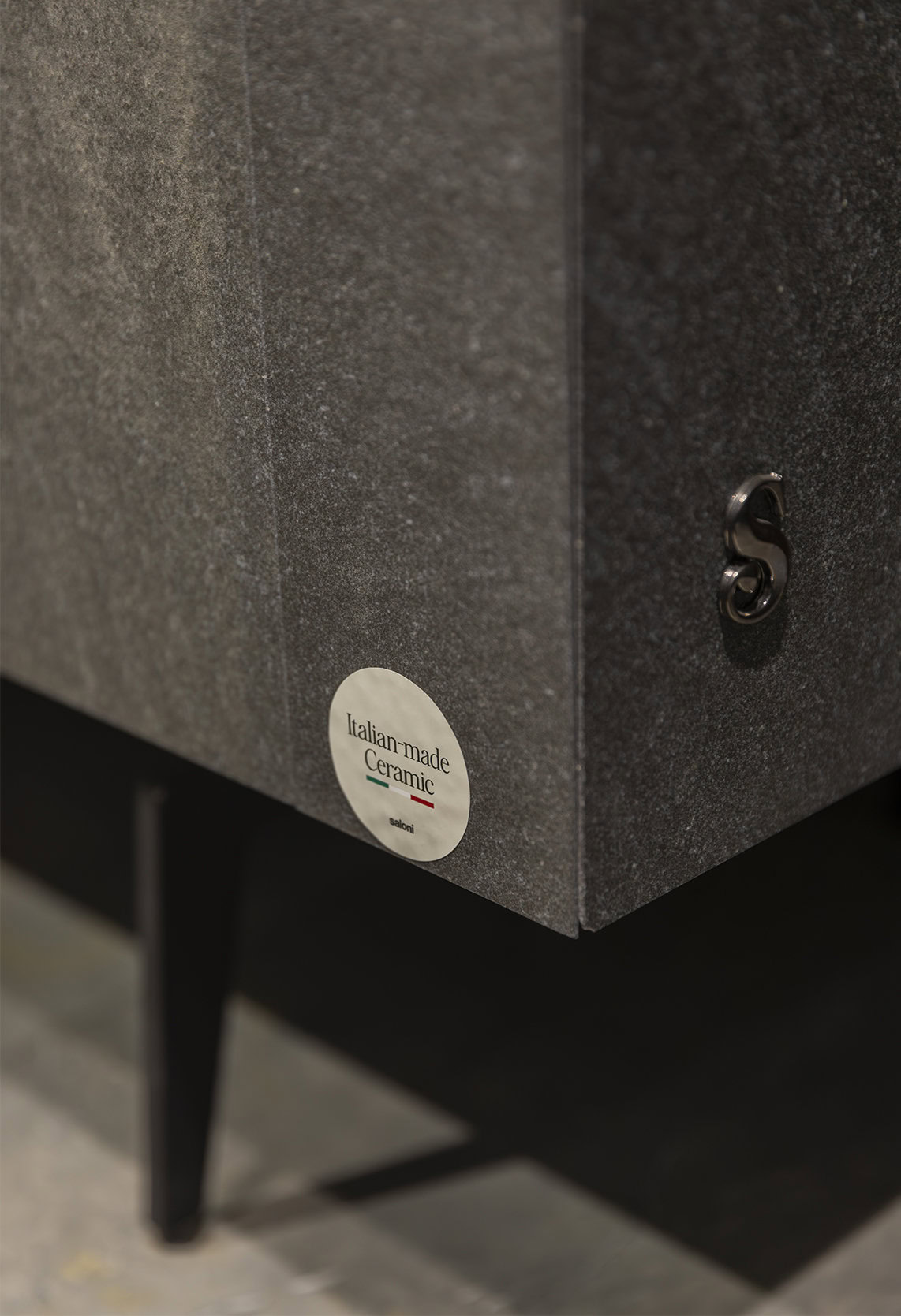 Sleek close-up of a Saloni Furniture handcrafted ceramic piece with Italian-made craftsmanship, featuring a textured dark gray surface and a small, elegant metallic S-shaped detail.