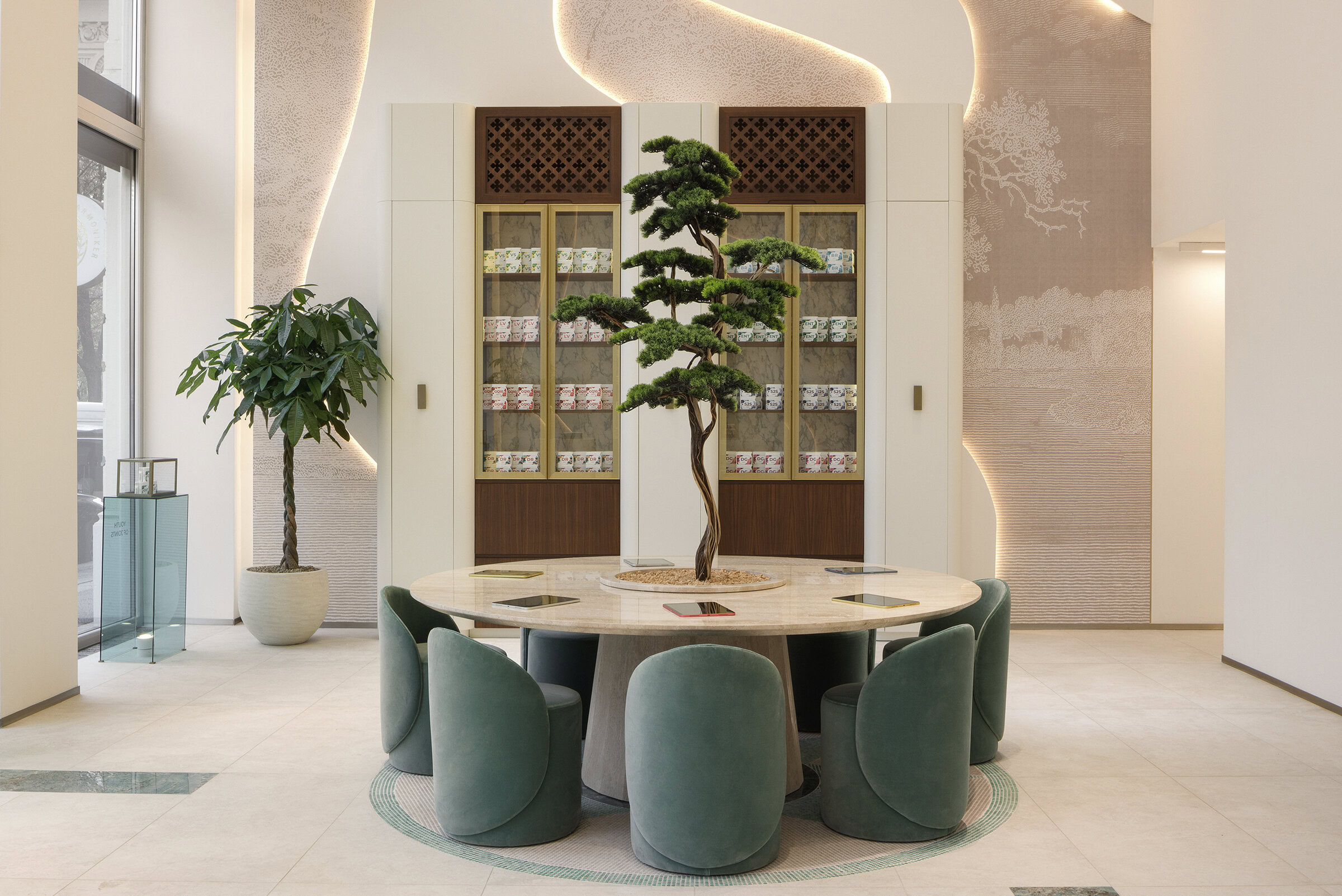 Stylish dining room featuring a round table with green velvet chairs, a central bonsai tree, and a contemporary bookshelf with decorative items in a well-lit space.