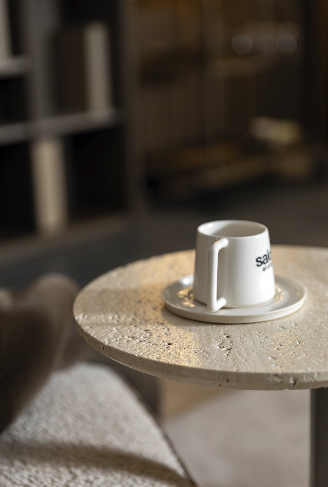 White ceramic coffee mug with Saloni Furniture logo on a round textured table in a cozy interior setting.